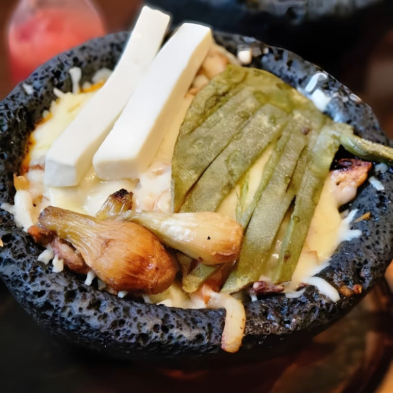 Molcajete dish with melted cheese, grilled nopales, and spring onions in a dark stone bowl.