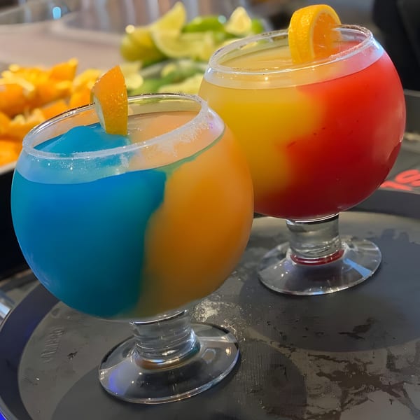 Two colorful El Agave Mexican Grill margaritas, one blue and orange, the other yellow and red, garnished with orange slices.