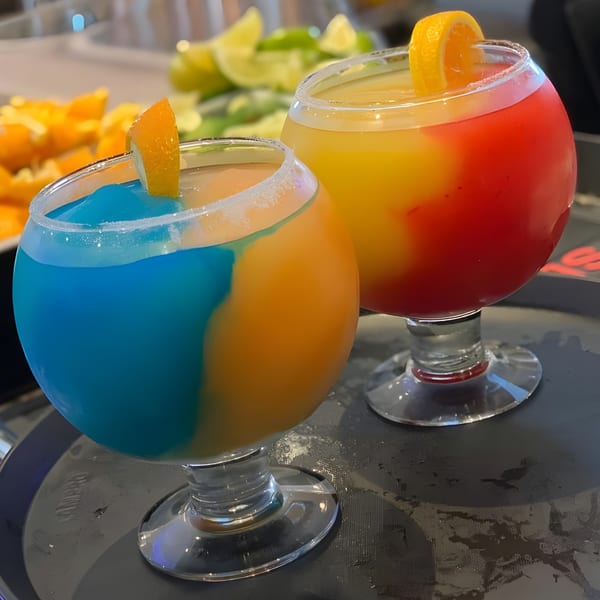 Two vibrant, multi-colored frozen margaritas with orange slices on a tray at El Agave Mexican Grill.